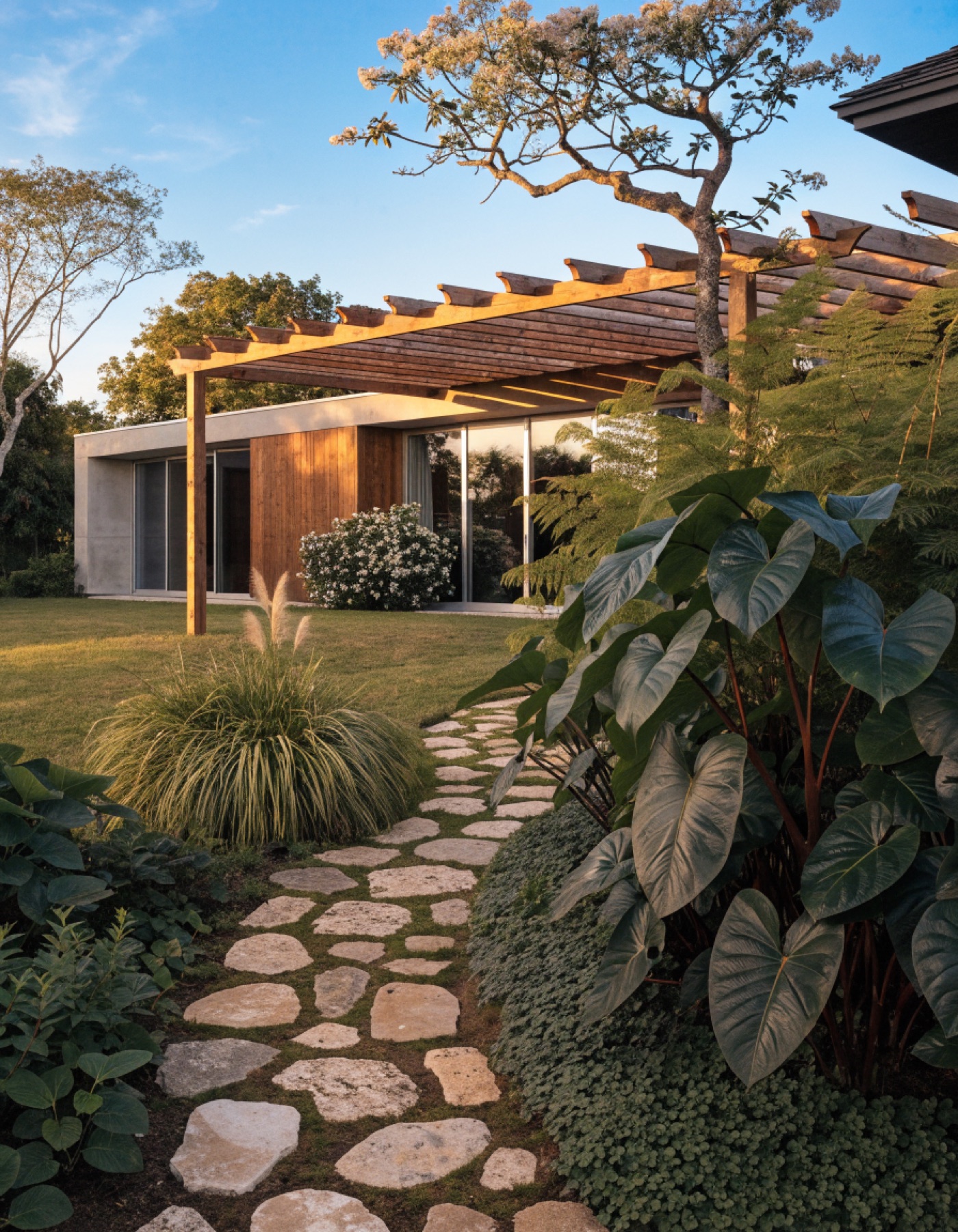 Lối đi đá tự nhiên dẫn vào ngôi nhà có pergola gỗ, hai bên là cây lá lớn và cỏ trang trí